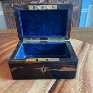 May include: Open antique wooden box with a dark, striped exterior and brass hardware. The interior is lined with blue velvet. The box is sitting on a wooden surface.