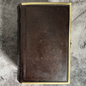 May include: A brown leather-bound book with a gold trim around the edges. The book is closed and lying flat on a gray surface.