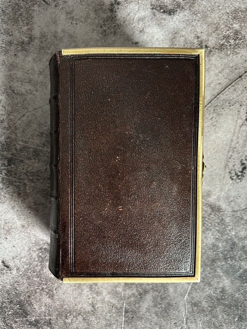 May include: A brown leather-bound book with a gold trim around the edges. The book is closed and lying flat on a gray surface.