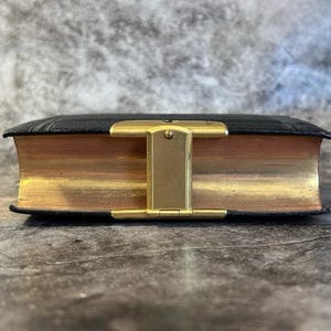 May include: A small, antique book with black leather covers and gold-gilded edges. The book is closed and secured with a gold-colored metal clasp. The pages are aged, giving the book a vintage appearance.