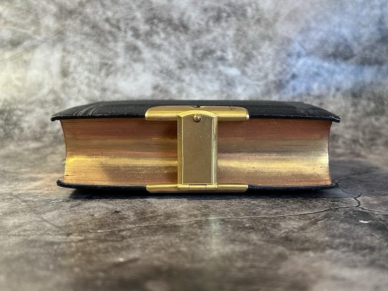 May include: A small, antique book with black leather covers and gold-gilded edges. The book is closed and secured with a gold-colored metal clasp. The pages are aged, giving the book a vintage appearance.