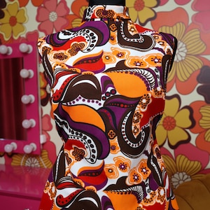 Könnte beinhalten: Ein ärmelloses Oberteil mit Stehkragen und einem lebendigen, mehrfarbigen abstrakten Blumen- und Paisley-Muster in Orange, Dunkellila, Braun, Rot und Weiß. Das Kleidungsstück hat eine strukturierte Stoffoberfläche und eine taillierte Passform.