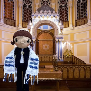 May include: Crocheted doll wearing a black outfit, a white and blue striped shawl, and a Star of David pendant. The doll is standing in a synagogue with stained glass windows and a golden ark.
