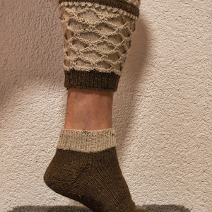 May include: Knitted leg warmers and socks in shades of brown and beige. The leg warmers feature a decorative openwork pattern and a brown band. The socks have a contrasting beige cuff. The items are displayed on a leg.