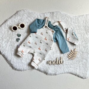 Op de afbeelding: Een baby-outfitset met een witte romper met dinosaurusprint, een lichtblauw shirt met lange mouwen en een bijpassende slab. De set bevat bloemvormige zonnebrillen en houten decoraties. De hashtag #ootd is ook zichtbaar.