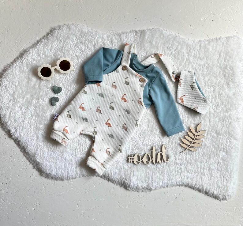 Op de afbeelding: Een baby-outfitset met een witte romper met dinosaurusprint, een lichtblauw shirt met lange mouwen en een bijpassende slab. De set bevat bloemvormige zonnebrillen en houten decoraties. De hashtag #ootd is ook zichtbaar.
