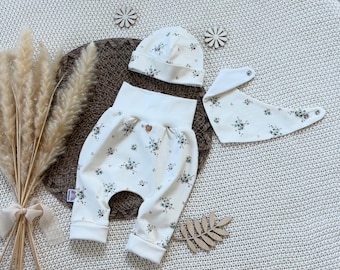 Op de afbeelding: Een babykledingset bestaande uit een off-white broek met bloemenpatroon, een bijpassende muts en een driehoekige slabbetje met drukknopen. De broek heeft een hoge tailleband en een klein houten knoopdetail in het midden aan de voorzijde.