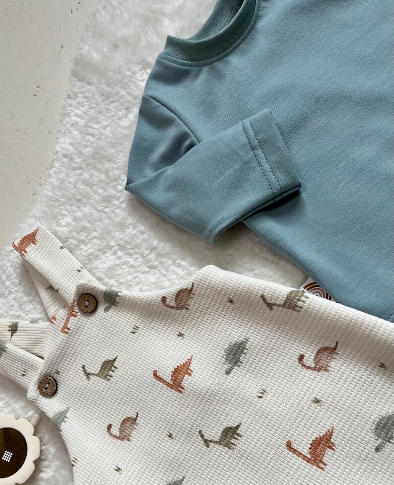 Op de afbeelding: Een lichtblauw shirt met lange mouwen en een witte wafel gebreide romper met een dinosauruspatroon. De romper heeft houten knopen en verstelbare bandjes. De kleding is ontworpen voor baby's en peuters.