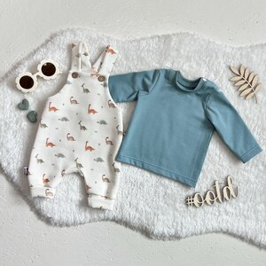 Op de afbeelding: Een baby-outfitset met een witte romper met dinosauruspatroon, een blauw shirt met lange mouwen en zonnebrillen in de vorm van bloemen. De set bevat een houten hashtag met de tekst #ootd.