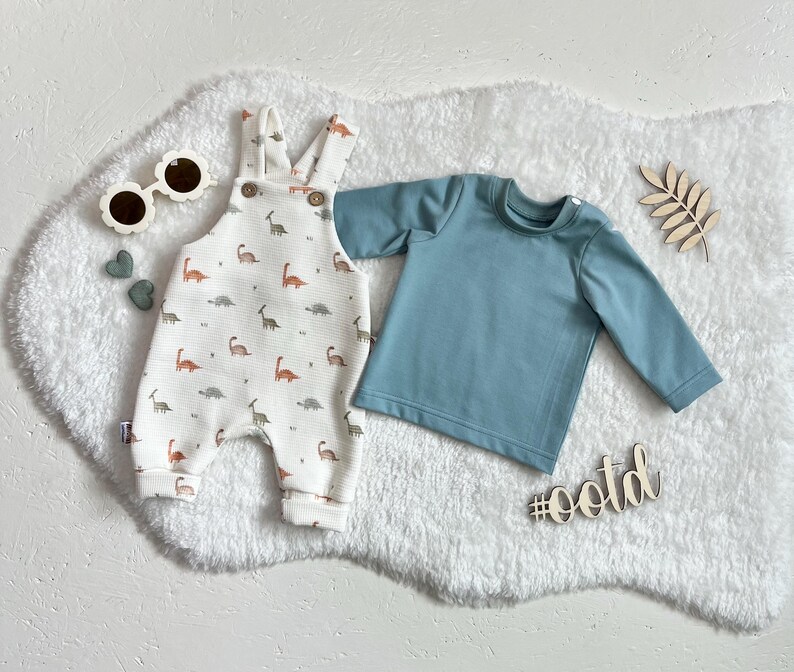 Op de afbeelding: Een baby-outfitset met een witte romper met dinosauruspatroon, een blauw shirt met lange mouwen en zonnebrillen in de vorm van bloemen. De set bevat een houten hashtag met de tekst #ootd.