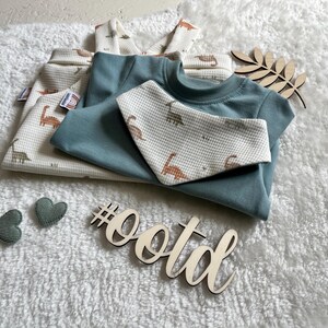 Op de afbeelding: Een set babykleding, waaronder een tealkleurig T-shirt en een slab, samen met een wafelgebreid shirt en broek met een dinosauruspatroon. Decoratieve houten elementen en hartvormige accenten zijn ook aanwezig. De hashtag #ootd is zichtbaar.
