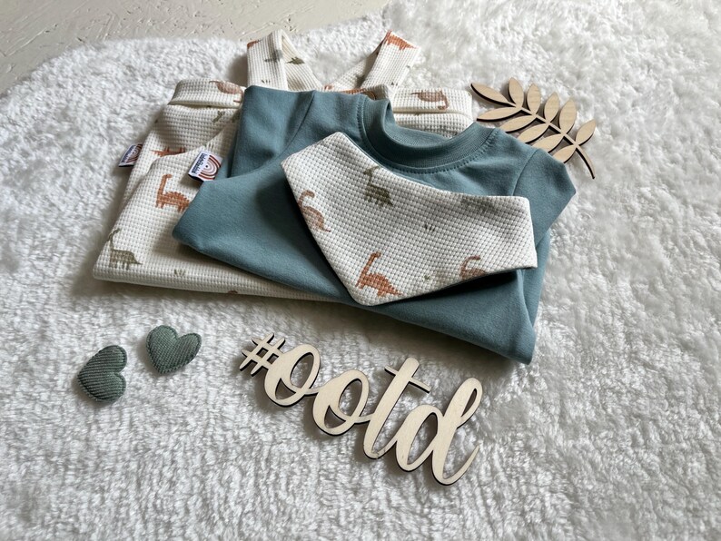 Op de afbeelding: Een set babykleding, waaronder een tealkleurig T-shirt en een slab, samen met een wafelgebreid shirt en broek met een dinosauruspatroon. Decoratieve houten elementen en hartvormige accenten zijn ook aanwezig. De hashtag #ootd is zichtbaar.