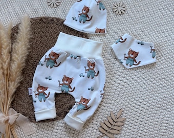 Op de afbeelding: Een witte babykledingset bestaande uit een broek met een hoge tailleband, een mutsje en een driehoekige slabbetje. De stof heeft een herhalend patroon van bruine tijgers in blauwe tuinbroeken met moersleutels en kleine blauwe speelgoedauto's.
