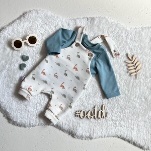 Op de afbeelding: Een baby-outfitset met een lichtblauw shirt met lange mouwen en een witte romper met een dinosauruspatroon. De romper heeft bretels en houten knopen. Accessoires: zonnebril en decoratieve houten vormen.