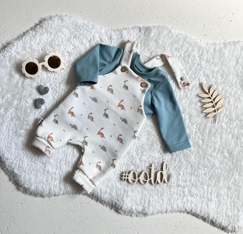 Op de afbeelding: Een baby-outfitset met een lichtblauw shirt met lange mouwen en een witte romper met een dinosauruspatroon. De romper heeft bretels en houten knopen. Accessoires: zonnebril en decoratieve houten vormen.