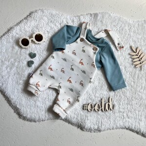 Op de afbeelding: Een baby-outfitset met een witte romper met een dinosauruspatroon en een blauw shirt met lange mouwen. De romper heeft houten knopen en bretels. De afbeelding bevat ook een zonnebril, hartvormige decoraties en een houten hashtag met #ootd.