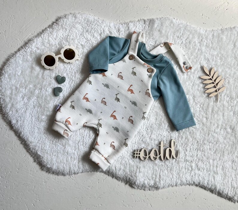 Op de afbeelding: Een baby-outfitset met een witte romper met een dinosauruspatroon en een blauw shirt met lange mouwen. De romper heeft houten knopen en bretels. De afbeelding bevat ook een zonnebril, hartvormige decoraties en een houten hashtag met #ootd.
