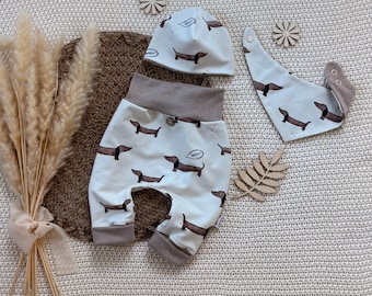 Op de afbeelding: Een babykledingset bestaande uit een mutsje, een broekje en een driehoekig slabbetje. Elk item is gebroken wit met een bruine teckelprint en de tekst "WOOF". Het broekje heeft een brede bruine tailleband en boorden aan de onderkant.