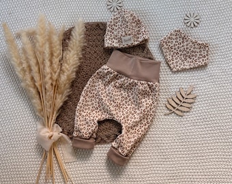 Op de afbeelding: Een babykledingset bestaande uit een mutsje met luipaardprint en een klein wit label met de tekst loved, een bijpassende bandana-slabbetje en een broekje met luipaardprint, beige boorden en een brede beige tailleband.