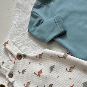 Op de afbeelding: Een lichtblauw shirt met lange mouwen en een witte wafel gebreide romper met een dinosauruspatroon. De romper heeft houten knopen en verstelbare bandjes. De dinosaurus illustraties zijn bruin en groen.