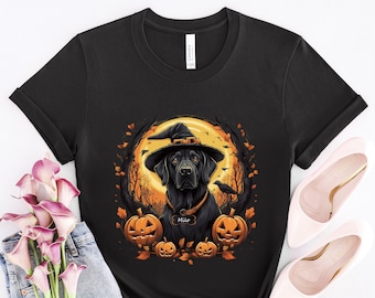 May include: Black Labrador Retriever wearing a witch hat and a collar with the name "Milo" on it. The dog is surrounded by pumpkins and fall leaves. The image is on a black t-shirt.