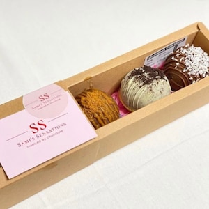 May include: A brown box with a pink label that says "Sami's Sensations Inspired by Chocolate" and the logo "SS". The box contains three chocolate bombs, one with a white chocolate coating and Oreo cookie pieces, one with a milk chocolate coating and coconut flakes, and one with a caramel coating and crushed graham crackers.