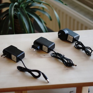 Pu&ograve; includere: Tre adattatori di alimentazione elettrici neri con cavi e spine su una superficie in legno chiaro.