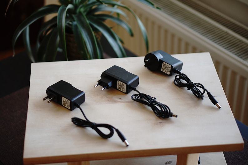 Pu&ograve; includere: Tre adattatori di alimentazione elettrici neri con cavi e spine su una superficie in legno chiaro.