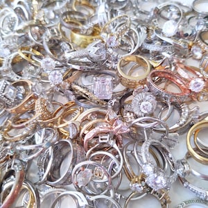 May include: A large pile of silver, gold, and rose gold rings with various styles and sizes. Some rings have clear gemstones, while others are plain bands.