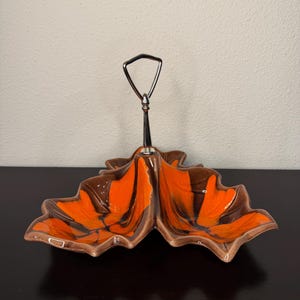 May include: A ceramic serving dish with a leaf-like design. The dish features a vibrant orange and brown color scheme with a silver handle. The dish is designed with two separate compartments, perfect for serving snacks or appetizers.