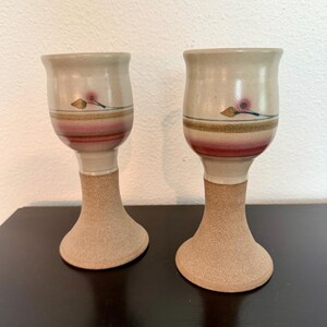 May include: Two handmade ceramic goblets. The goblets have a beige base with a textured, flared foot and stem. The upper portion is beige with a red and brown band and a floral design. The goblets are approximately 6 inches tall.