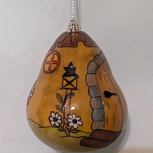 May include: Hand-painted gourd ornament shaped like a house. The ornament features a brown roof, a round window, and a wooden door. A black lantern and white daisies are painted on the front. A silver string is attached for hanging.