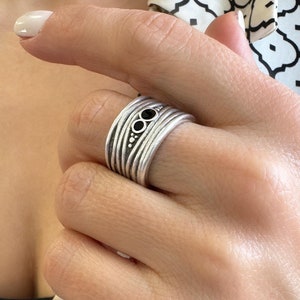 Op de afbeelding: Een zilveren ring met meerdere banden en drie zwarte edelstenen. De ring wordt aan een vinger gedragen.