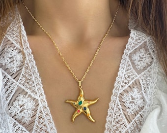 May include: A gold-toned necklace featuring a starfish-shaped pendant. The pendant is adorned with six small, multi-colored inset stones in shades of green, blue, red, and orange, suspended from a delicate gold-toned chain link necklace.