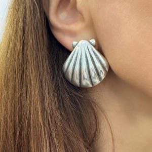 以下が含まれることがあります： 銀色の貝殻型のイヤリング。イヤリングは、リブのあるテクスチャを備えた詳細な3次元デザインで、耳に装着されています。イヤリングはスタッドスタイルで、クローズアップショットです。