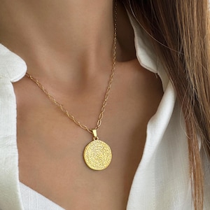 Puede incluir: Un collar de cadena de oro con un gran colgante redondo que presenta diseños intrincados y detallados. El colgante está hecho de metal dorado y tiene una forma circular.