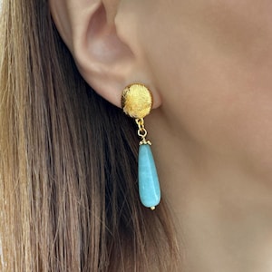 Könnte beinhalten: Goldfarbene Ohrringe mit einem strukturierten, kreisförmigen Stecker und einem tropfenförmigen, hellblauen Edelsteinanhänger. Die Ohrringe sind hängend und haben ein zartes Design, das eine Mischung aus Texturen und Farben zeigt.