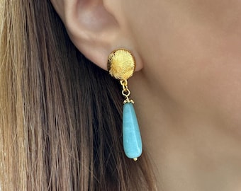 May include: Gold-colored earrings with a textured, circular stud and a teardrop-shaped, light blue gemstone pendant. The earrings are dangling and have a delicate design, showcasing a blend of textures and colors. The jewelry is worn on an ear.