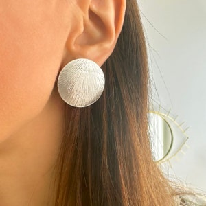 May include: Silver earrings with a textured, circular design. The earrings are large and have a slightly raised center.