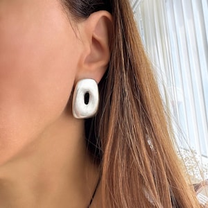 May include: A silver-coloured, rectangular stud earring with a rounded shape and a central oval cutout. The earring has a matte finish and is worn on the ear. The jewellery is a modern, minimalist design.