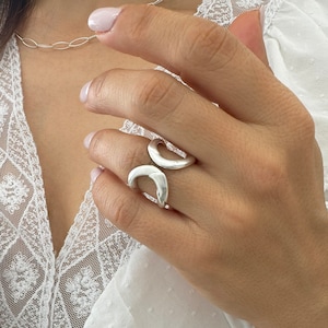 Peut inclure: Une bague en argent au design ouvert unique. La bague présente deux éléments incurvés en forme de croissant qui entourent le doigt. La bague est portée sur une main avec un haut blanc et un collier en chaîne argentée.