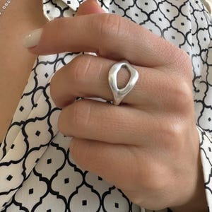 Op de afbeelding: Een zilveren ring met een ovale uitsparing wordt op een hand getoond. De ring heeft een glad, gepolijst oppervlak en een modern, minimalistisch ontwerp. De achtergrond omvat een zwart-witte blouse met patroon.