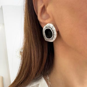 Può includere: Un orecchino a lobo color argento dalla forma organica irregolare, caratterizzato da un bordo testurizzato con linee radiali che circonda un inserto centrale nero, liscio e lucido. Il pezzo presenta una finitura metallica e un aspetto stratificato.
