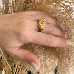 Może przedstawiać: Złoty pierścionek z wzorem w kształcie oka. Pierścionek jest ozdobiony okrągłym, wypukłym elementem centralnym i ma fakturowaną powierzchnię.