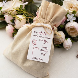 May include: A white fabric bag with a tag that reads "The World needs more women like You Happy Women's Day! XOXO, Pam". The tag has a red rose illustration.