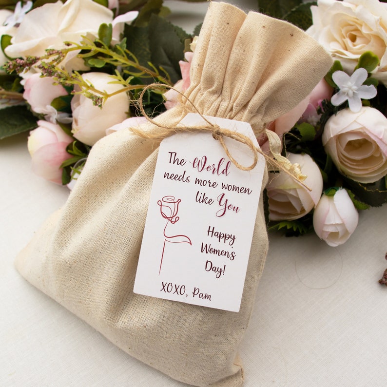 May include: A white fabric bag with a tag that reads "The World needs more women like You Happy Women's Day! XOXO, Pam". The tag has a red rose illustration.