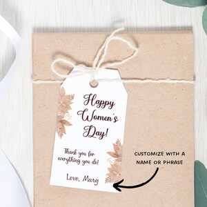 May include: A white gift tag with brown floral designs and the text "Happy Women's Day! Thank you for everything you do! Love, Mary". The tag is tied with twine to a brown paper gift bag. The text "CUSTOMIZE WITH A NAME OR PHRASE" is written on the bag.