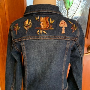 May include: A dark blue denim jacket with embroidered autumnal designs on the back. The designs include a squirrel, a mushroom, and leaves.