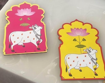 May include: Two decorative wooden plaques with a pink and yellow background. Each plaque features a white cow with black spots and a pink lotus flower.