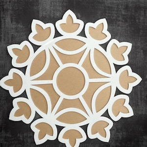 May include: A white paper mandala with a brown center. The mandala has a circular design with intricate details and a floral pattern around the edge.